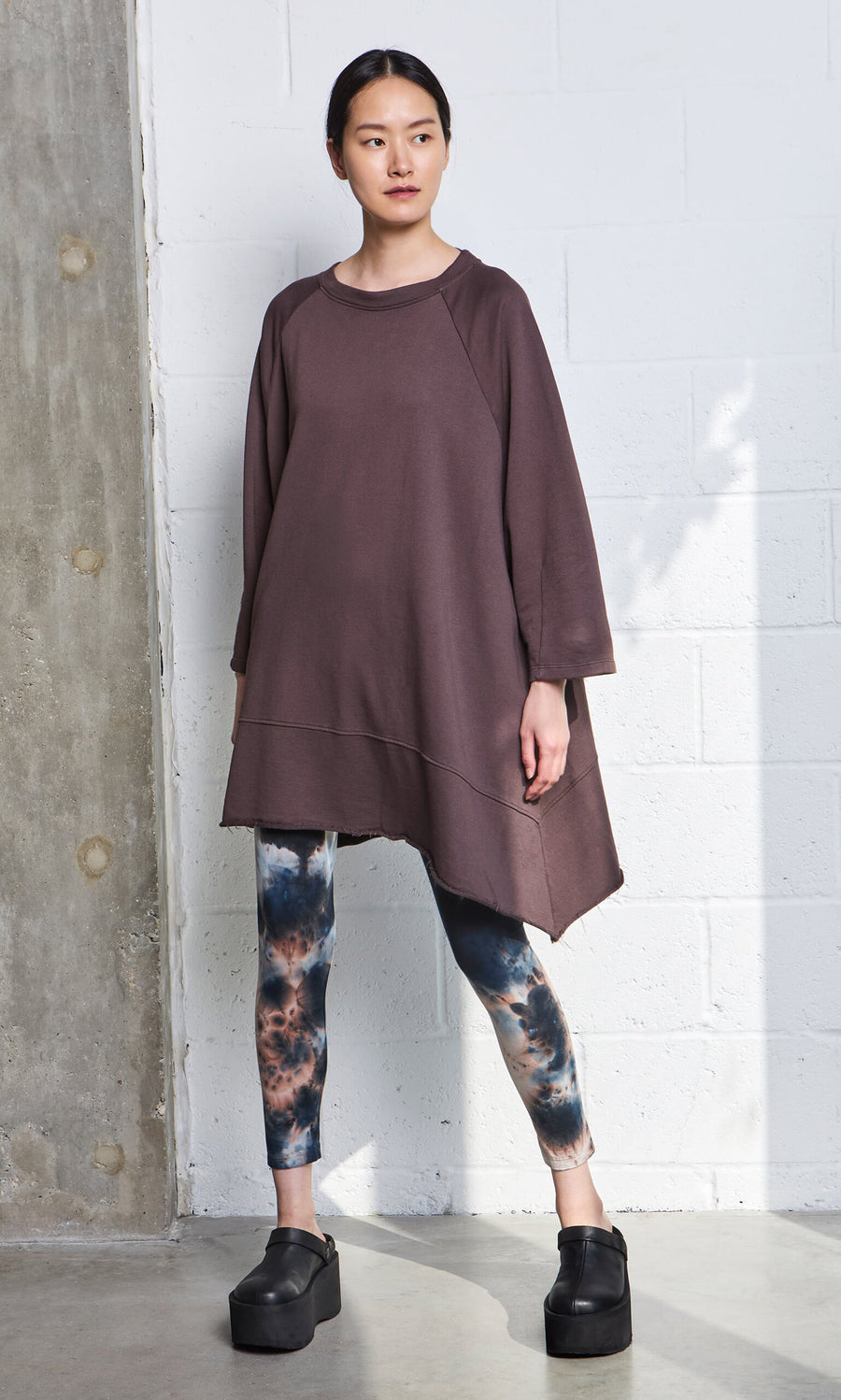 Plümo- Woman in Tie Dye Leggings and brown tunic with black platforms stands by a concrete and white brick wall.