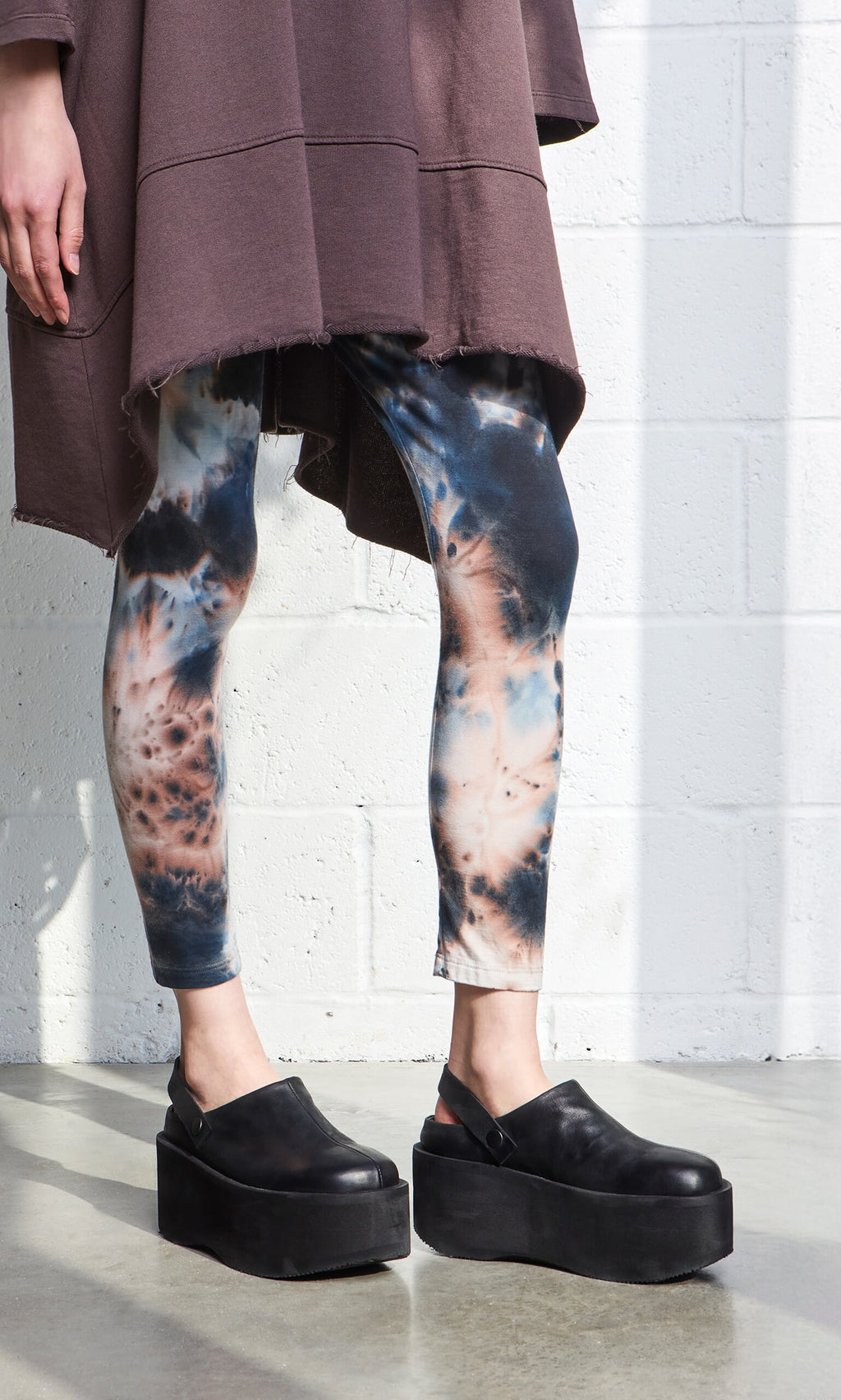 Plümo- Person in brown oversized top, Tie Dye Leggings, and black platform shoes stands by a white brick wall.