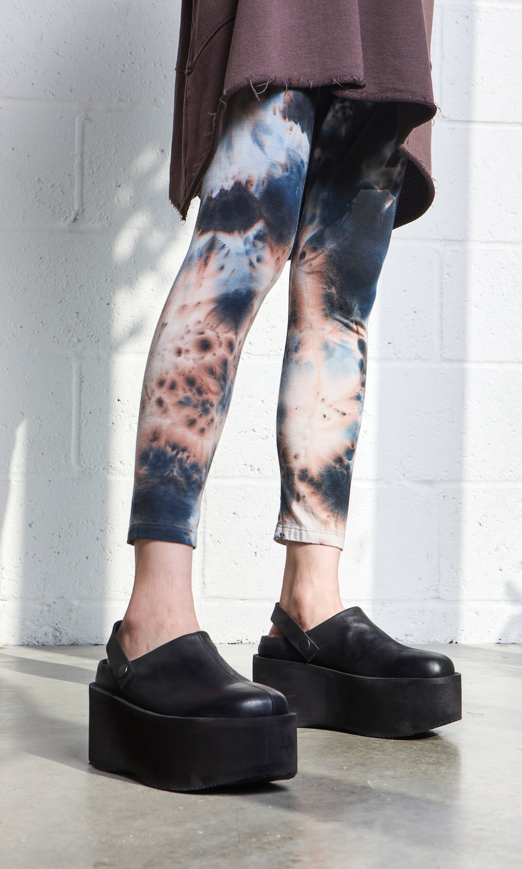 Plümo- Person in Tie Dye Leggings and black platform shoes, brown top, on a concrete floor by a white wall.