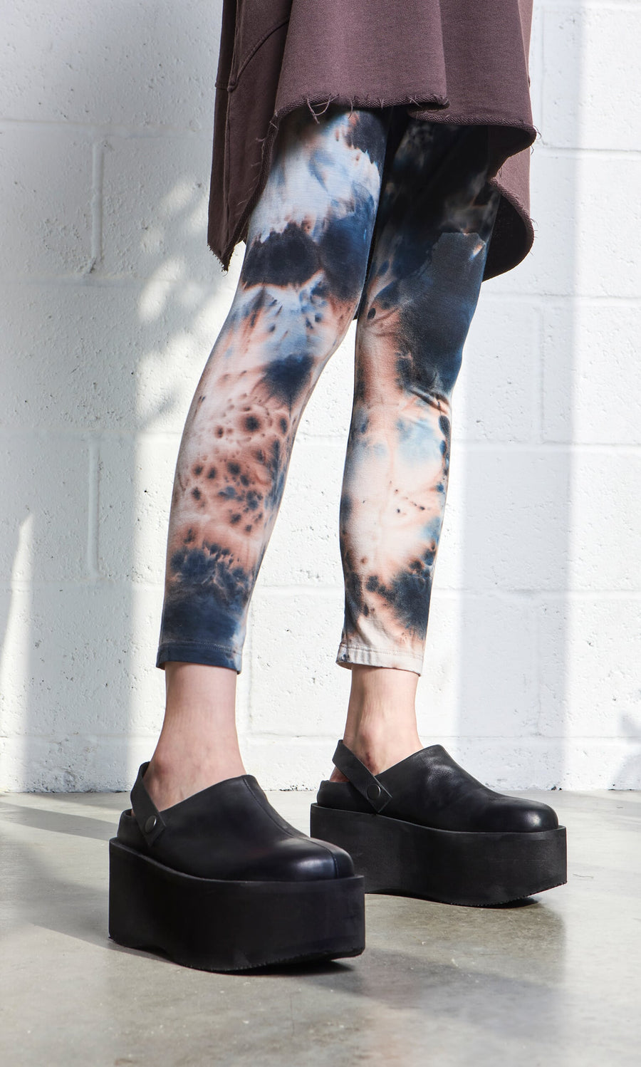 Plümo- Person in Tie Dye Leggings and black platform shoes, brown top, on a concrete floor by a white wall.