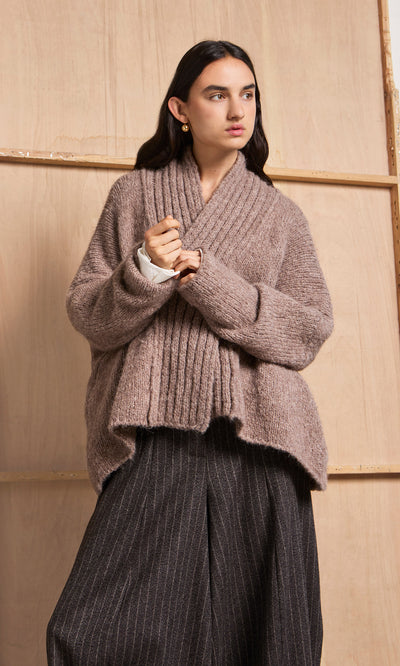 Plümo  Person in a Shawl Collar Jumper and wide pinstripe pants stands before a wooden background.