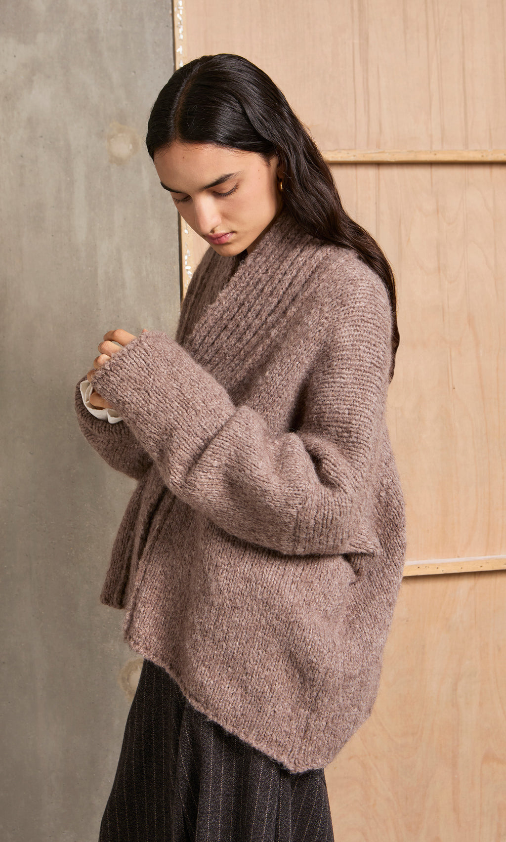 Plümo  Woman in a Shawl Collar Jumper with chunky ribbed knit adjusts her sleeve while standing indoors.