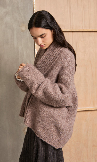 Plümo  Woman in a Shawl Collar Jumper with chunky ribbed knit adjusts her sleeve while standing indoors.