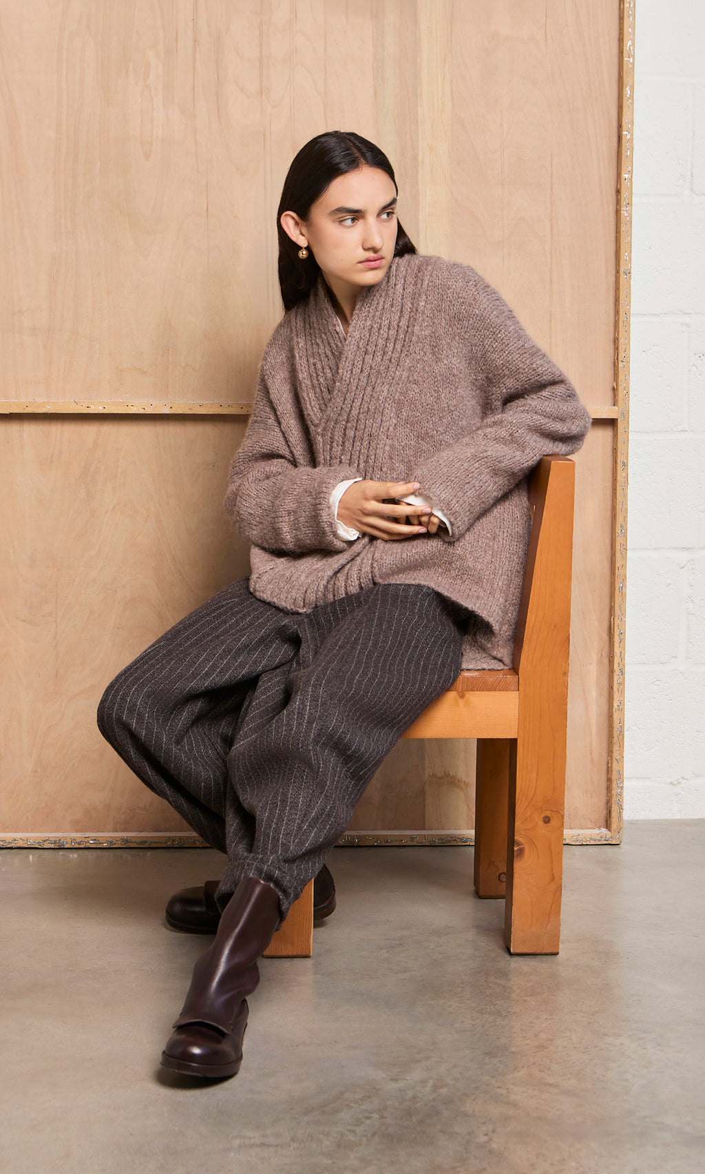 Plümo  A woman in a Shawl Collar Jumper and pinstripe pants sits on a wooden chair, looking left with hands clasped.