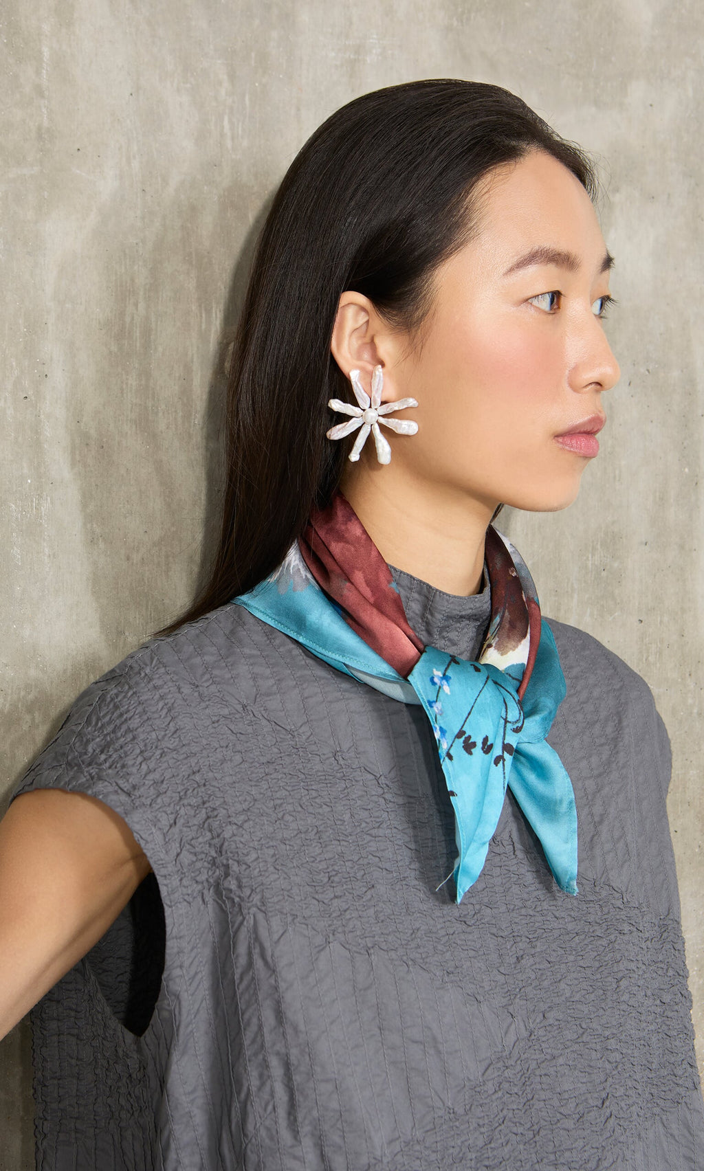 Plümo- Woman with long dark hair wears an Ultramarine Necktie, gray textured top, and floral earrings, facing sideways.