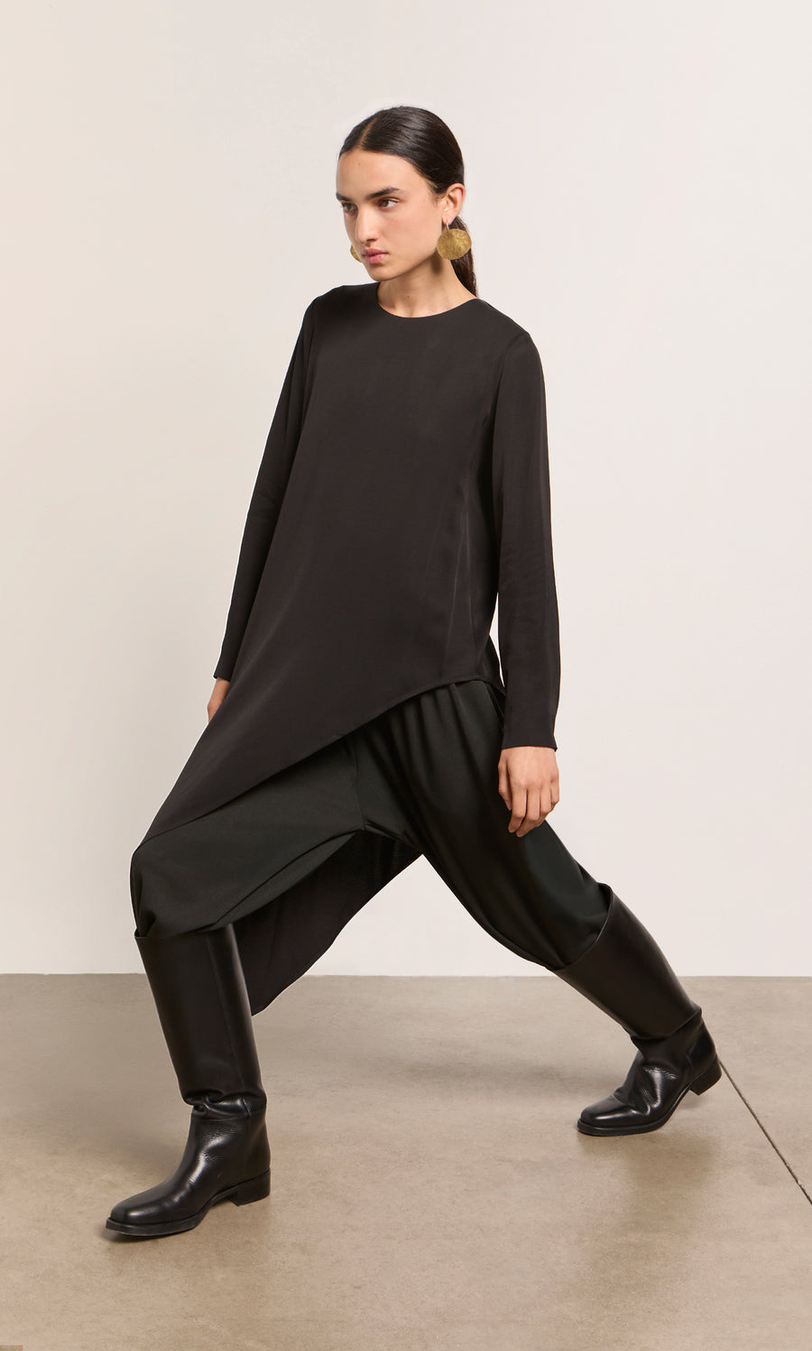 Plümo  Woman in Vermeer Top, wide pants, and boots poses with one leg bent forward against a plain background.