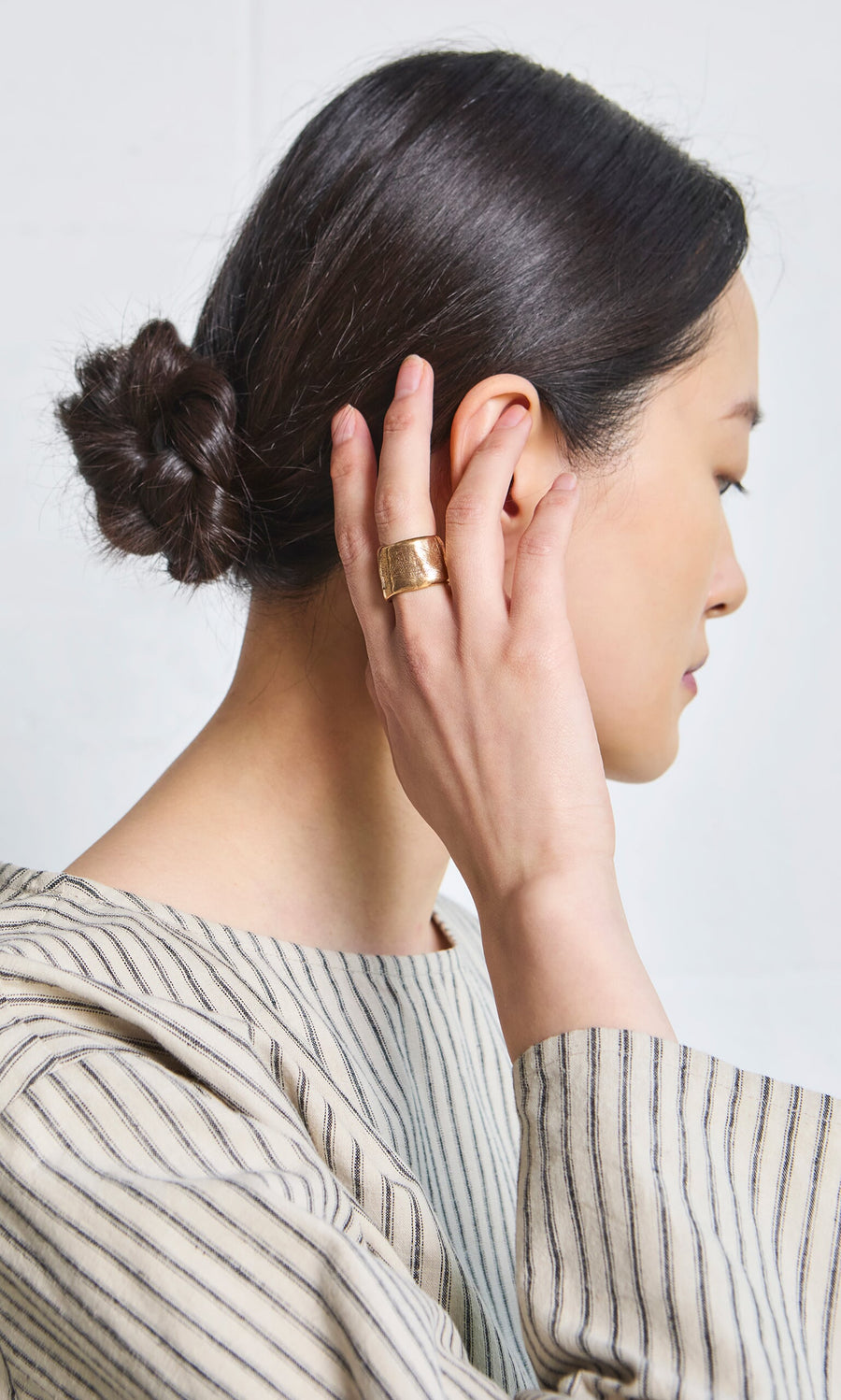 Plümo- Woman with dark hair in a low bun, striped top, wearing the chunky handmade Vinci Ring, touching her ear.