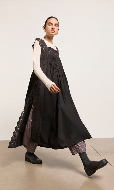 Plümo  Person in a Vintage Lace Dress over striped pants and black shoes walks indoors, showing maxi pinafore style.