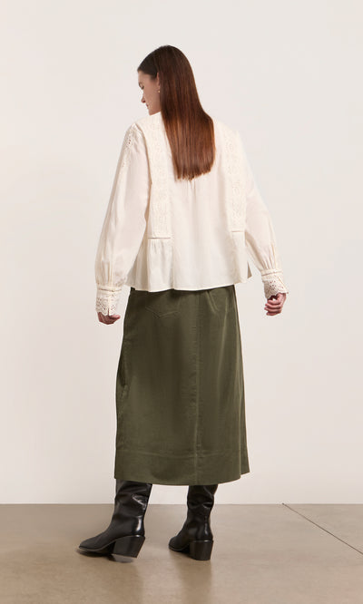 Plümo  Woman in a Beetle Green Skirt, white blouse, and black boots stands against a plain light background, facing away.