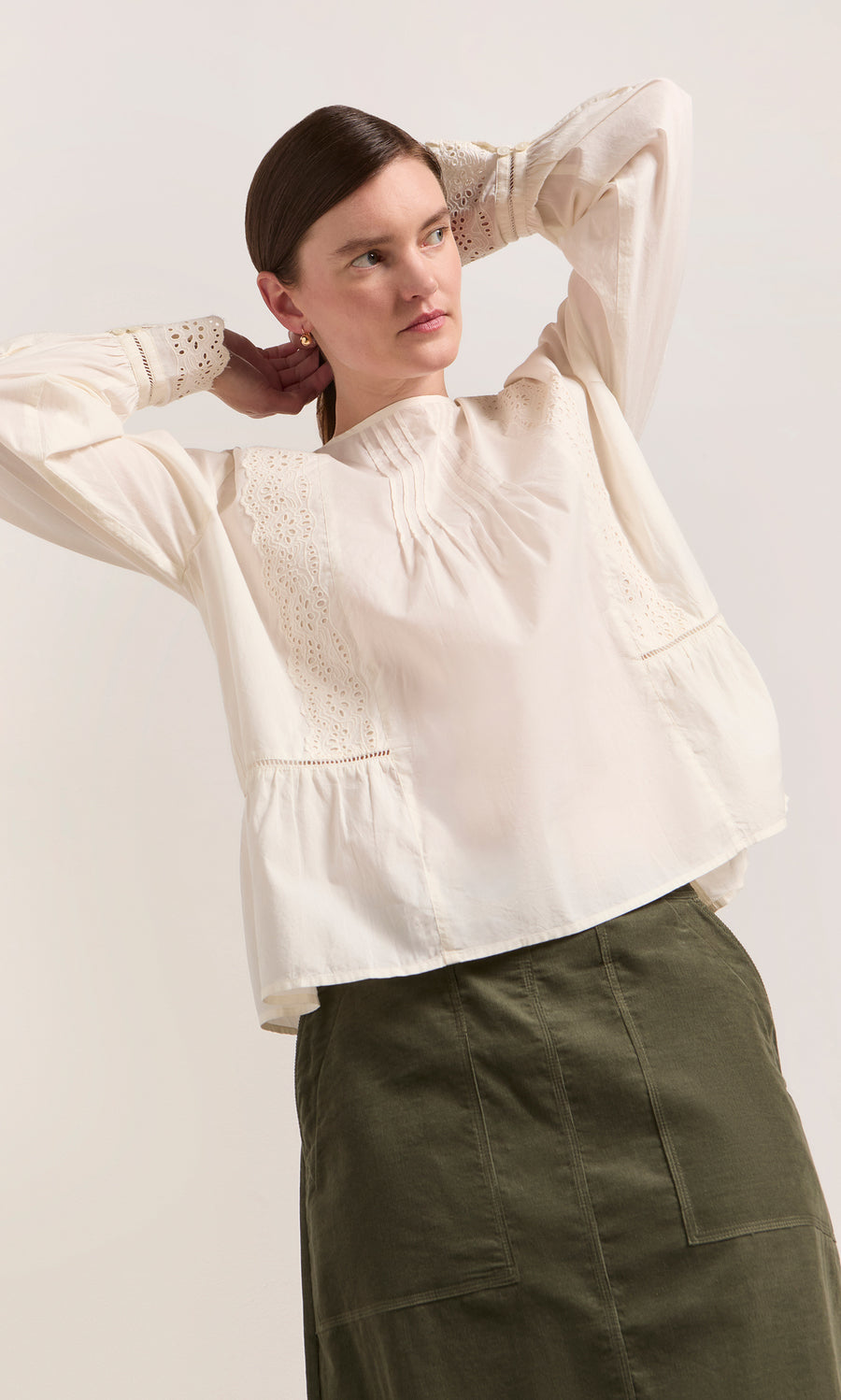 Plümo  Woman in a White Broderie Shirt and olive skirt stands with hands behind her head, gazing to the side.