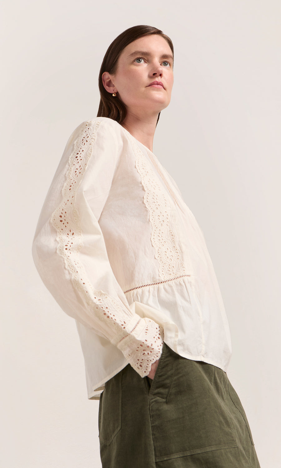 Plümo  Woman wears a White Broderie Shirt and olive green pants, standing against a plain background.