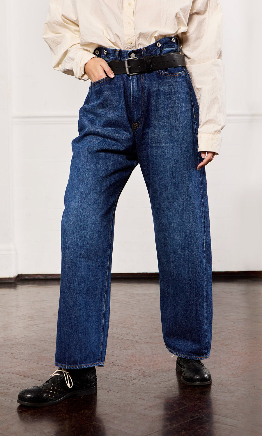 Plümo- Person wears Stack Jeans, a beige shirt, black belt, and black shoes while standing on a dark wood floor indoors.