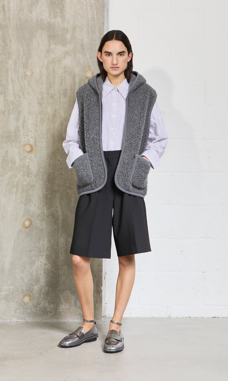 Plümo - Person stands by a concrete wall in a snug fit Wool Gilet - Graphite, striped shirt, black shorts, and metallic shoes.