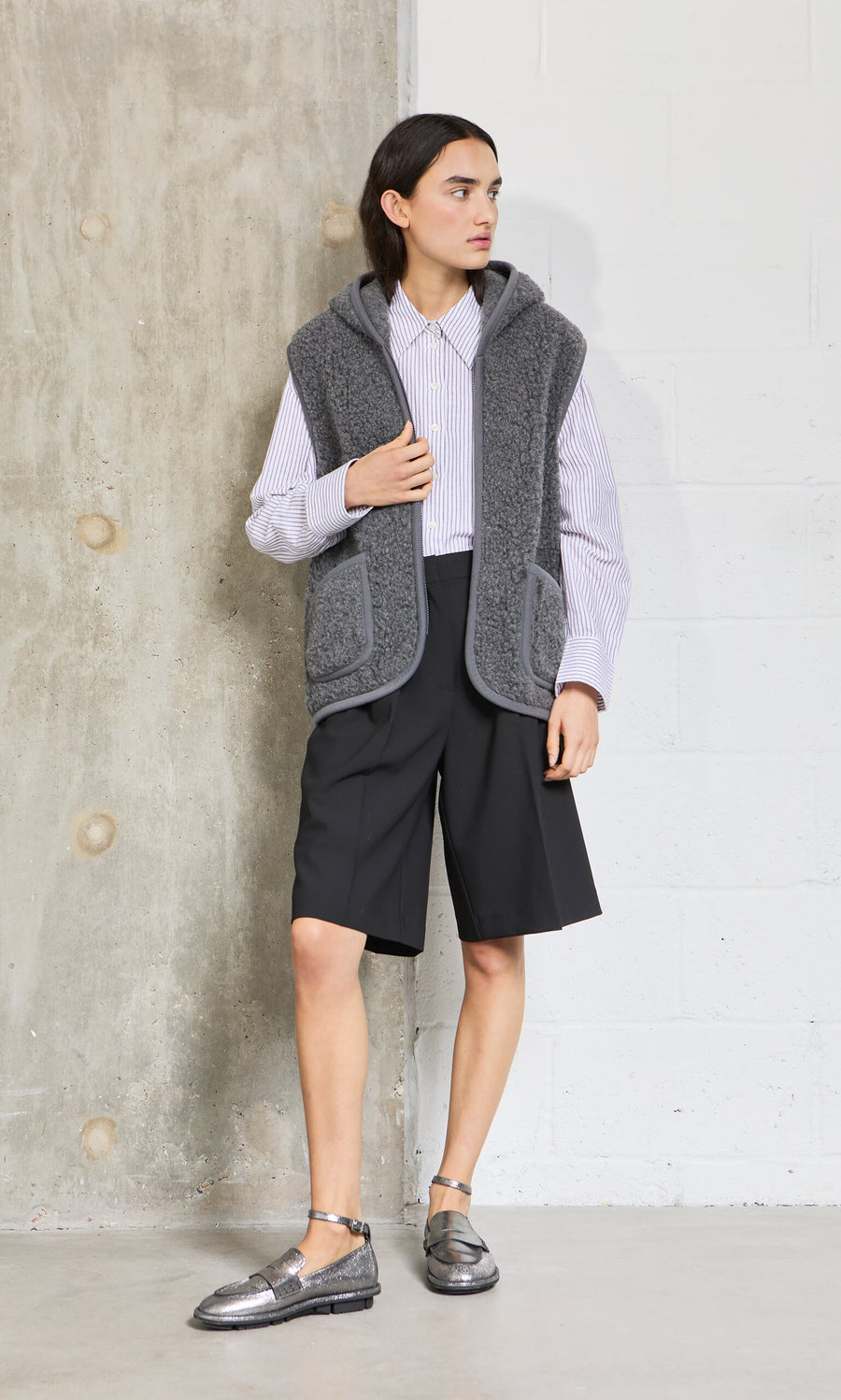 Person wearing a grey hooded vest over a white shirt and black shorts, standing against a concrete wall.