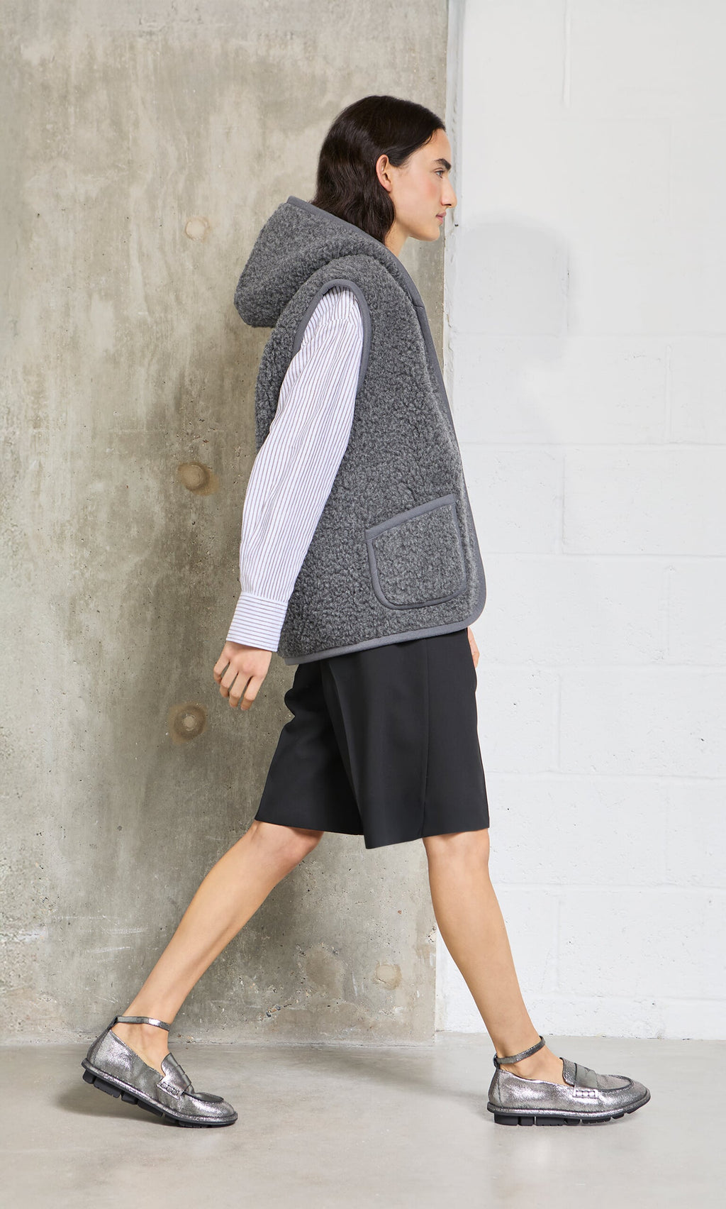 Plümo - Person in a graphite Wool Gilet, striped shirt, black shorts, and silver shoes walks indoors by a concrete wall.
