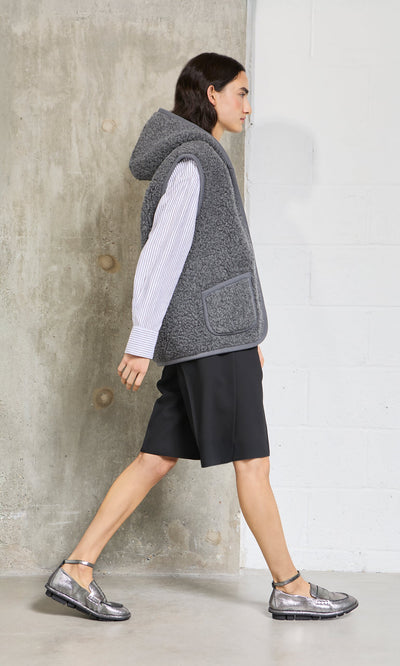Plümo - Person in a graphite Wool Gilet, striped shirt, black shorts, and silver shoes walks indoors by a concrete wall.