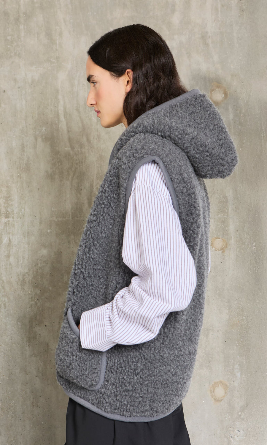 Person wearing a gray hooded vest over a white shirt against a concrete wall.