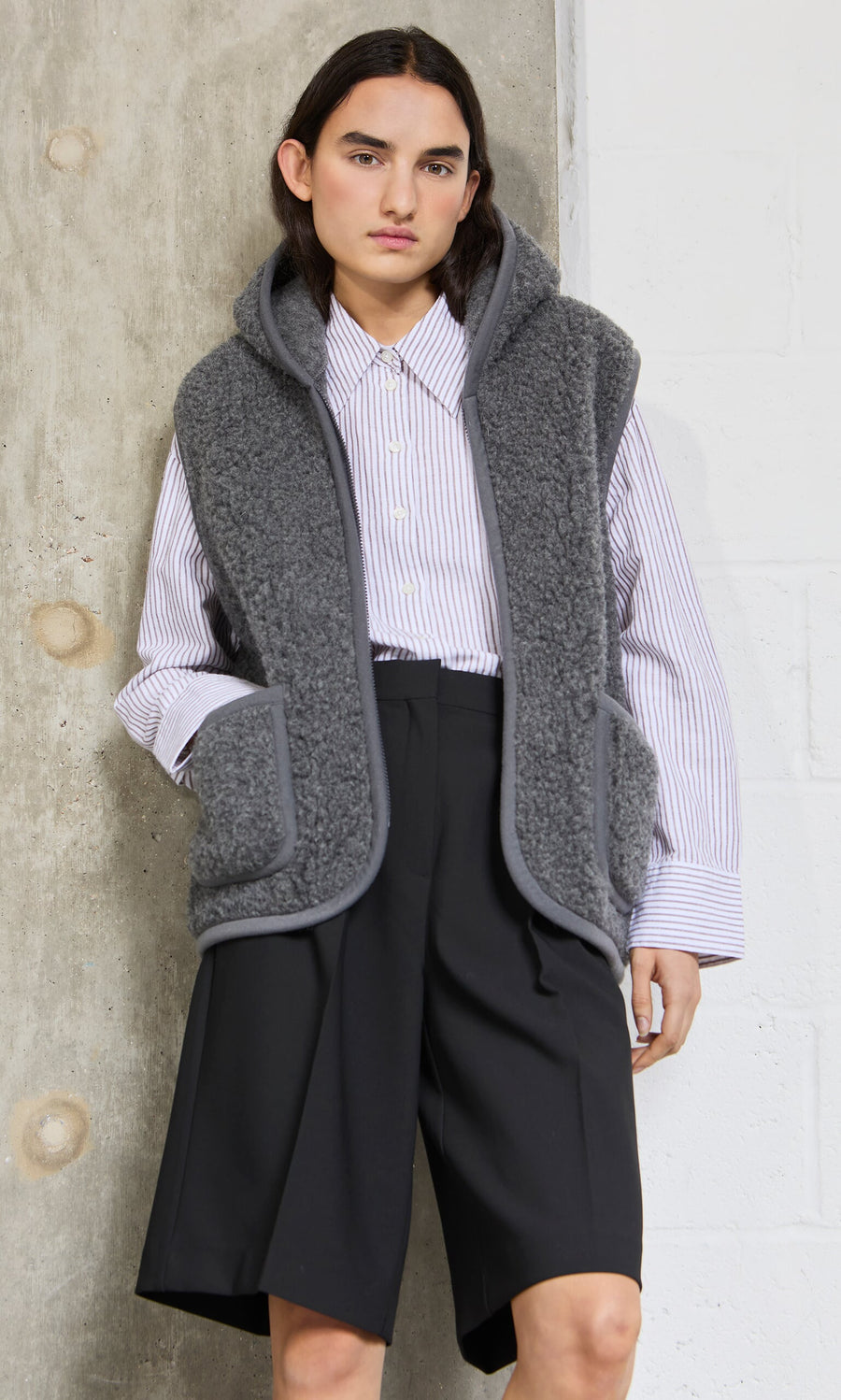 Plümo - Person wearing the Wool Gilet - Graphite over a striped shirt and black shorts, standing by a concrete wall.