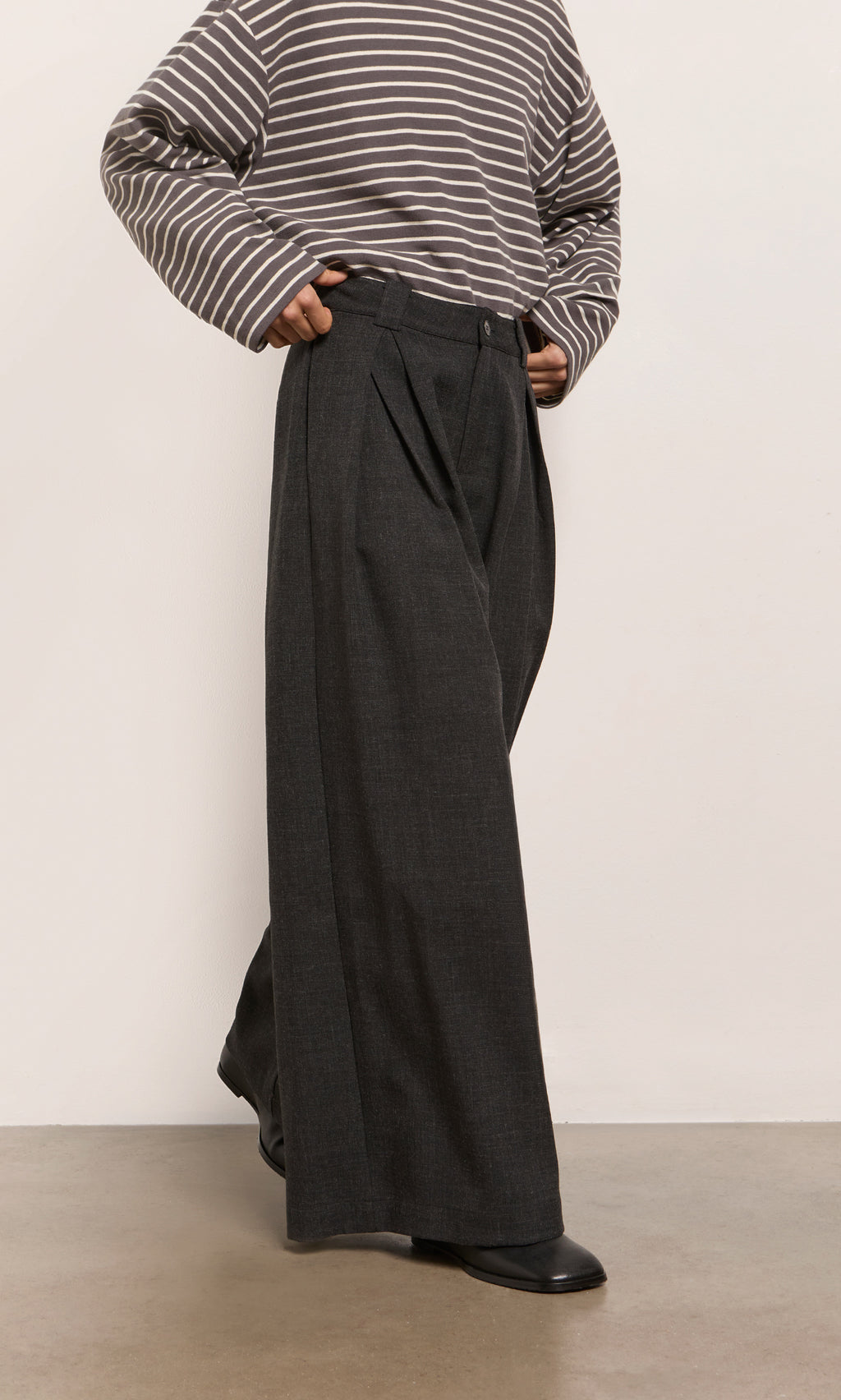 Plümo  Person in a striped shirt, Pleated Grey Pants, and black shoes stands on a light-colored floor.
