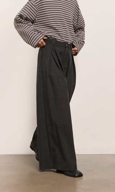 Plümo  Person in a striped shirt, Pleated Grey Pants, and black shoes stands on a light-colored floor.