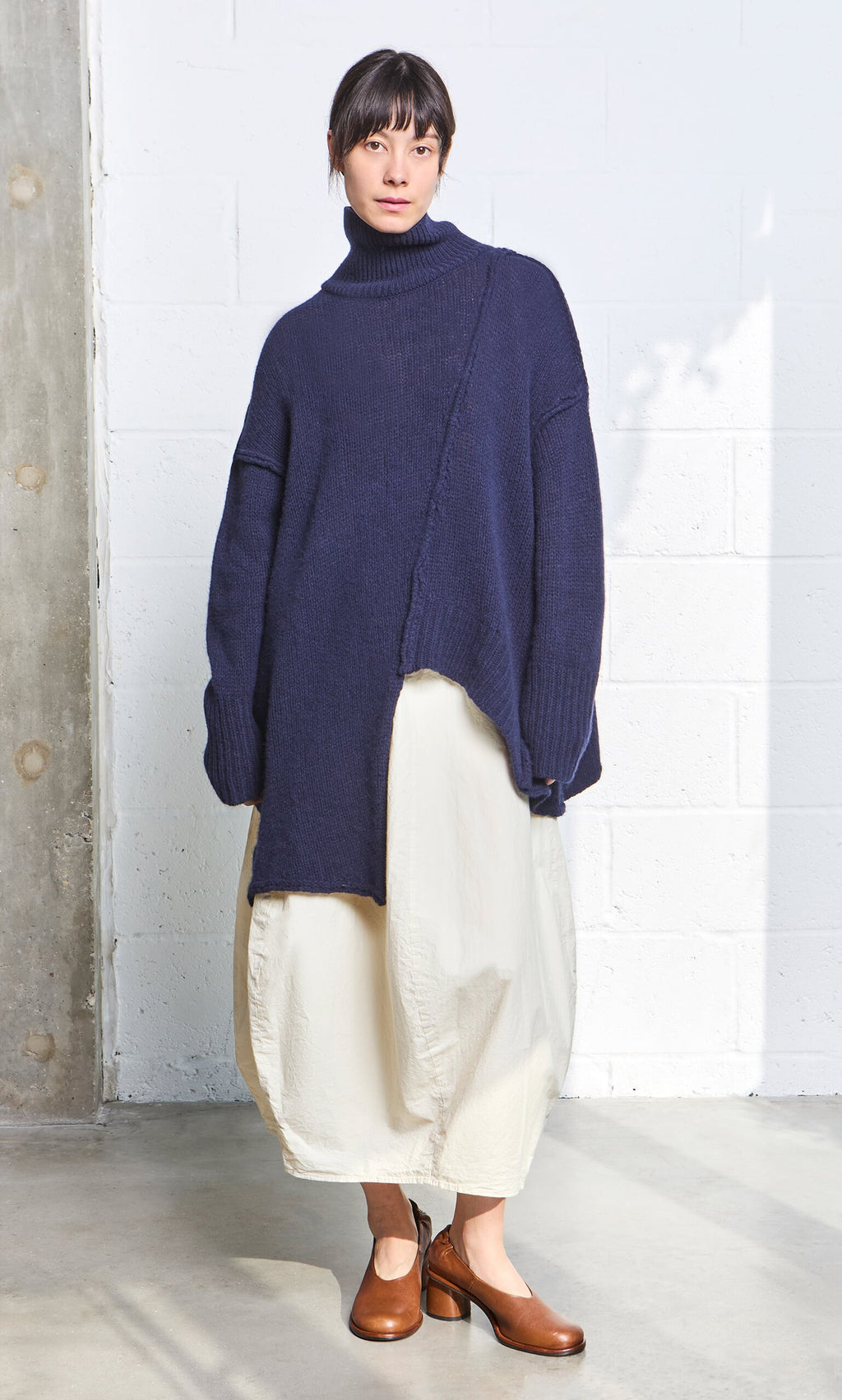 Plümo- Woman in a navy Yarker Rollneck sweater, white skirt, and brown heels stands by a white brick wall and pillar.