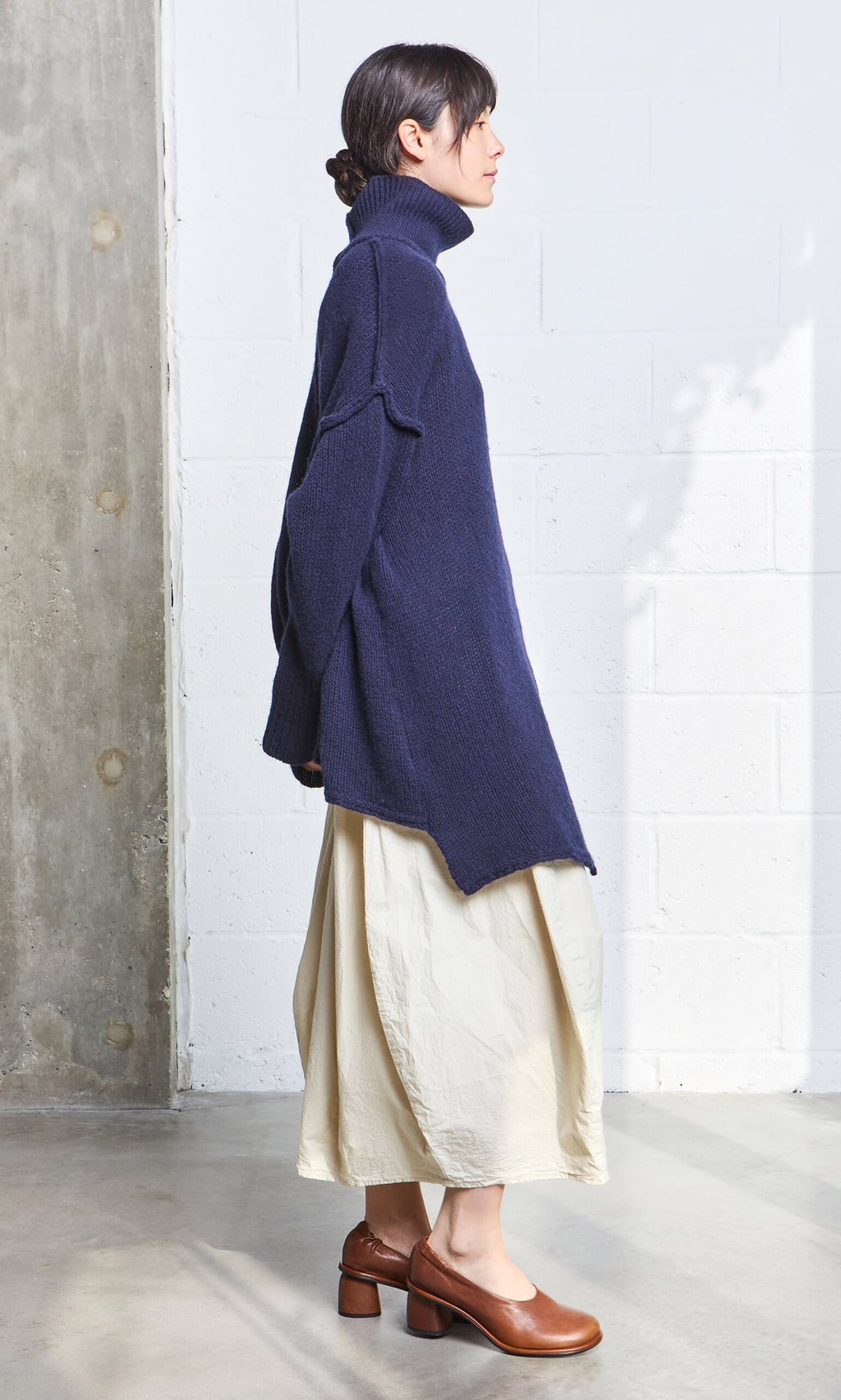 Plümo- Woman in a navy Yarker Rollneck, cream skirt, and brown heels stands sideways indoors.