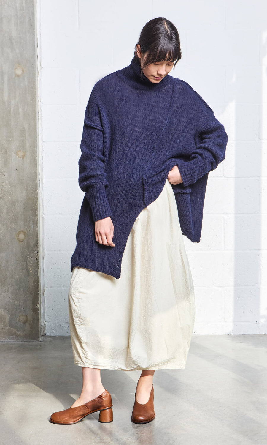 Plümo- Woman in a Yarker Rollneck navy sweater, cream skirt, and brown heels stands indoors on a concrete floor.