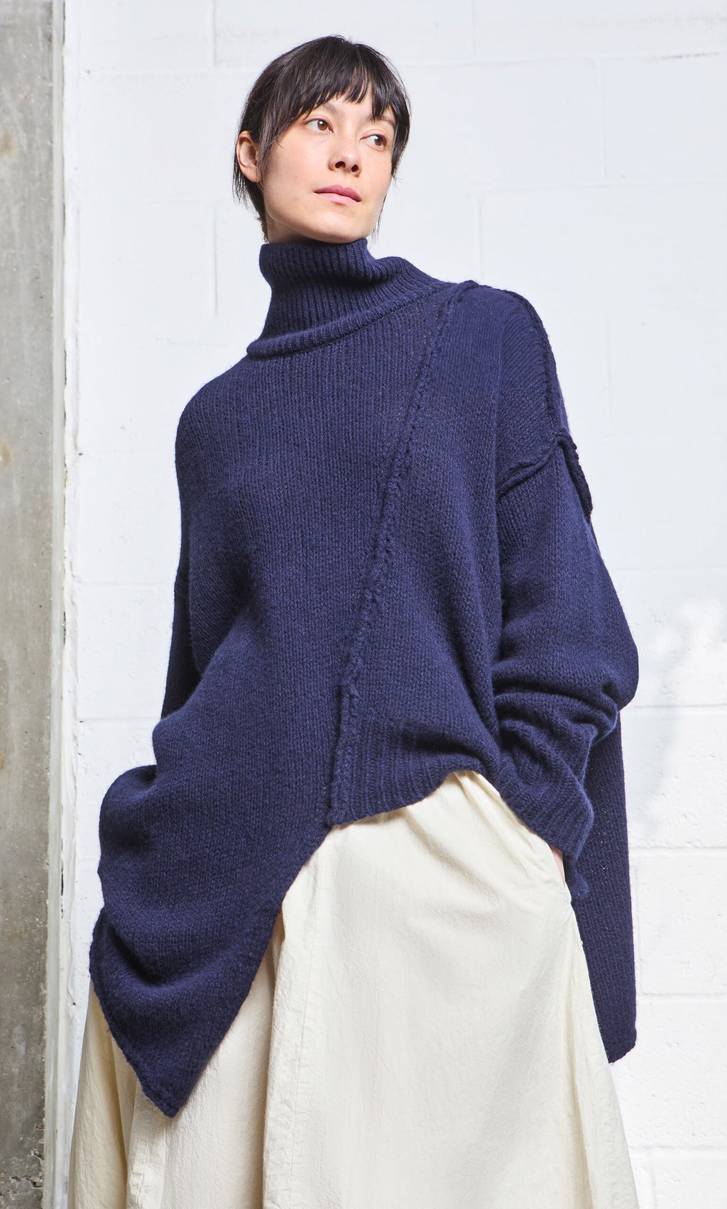 Plümo- Woman in a navy blue Yarker Rollneck sweater and off-white skirt, standing against a light-colored wall.