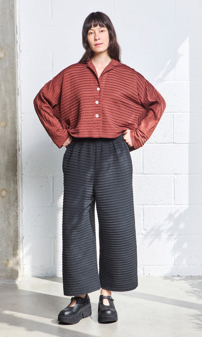 Plümo- Woman standing indoors in a rust button-up top and black Koji Pants with Japanese-inspired textured patterns.