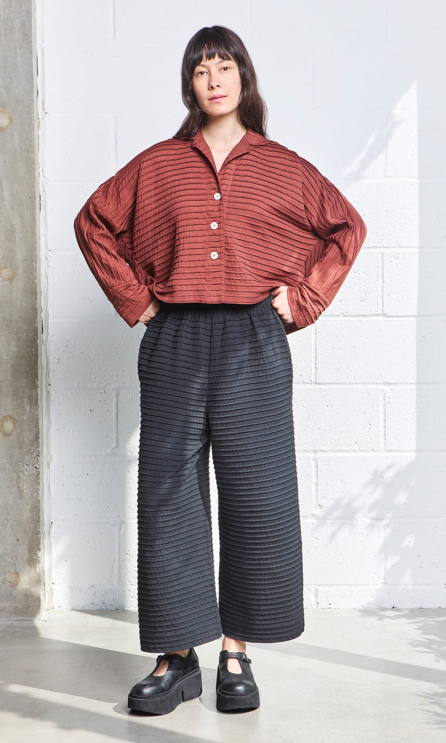 Plümo- Woman standing indoors in a rust button-up top and black Koji Pants with Japanese-inspired textured patterns.