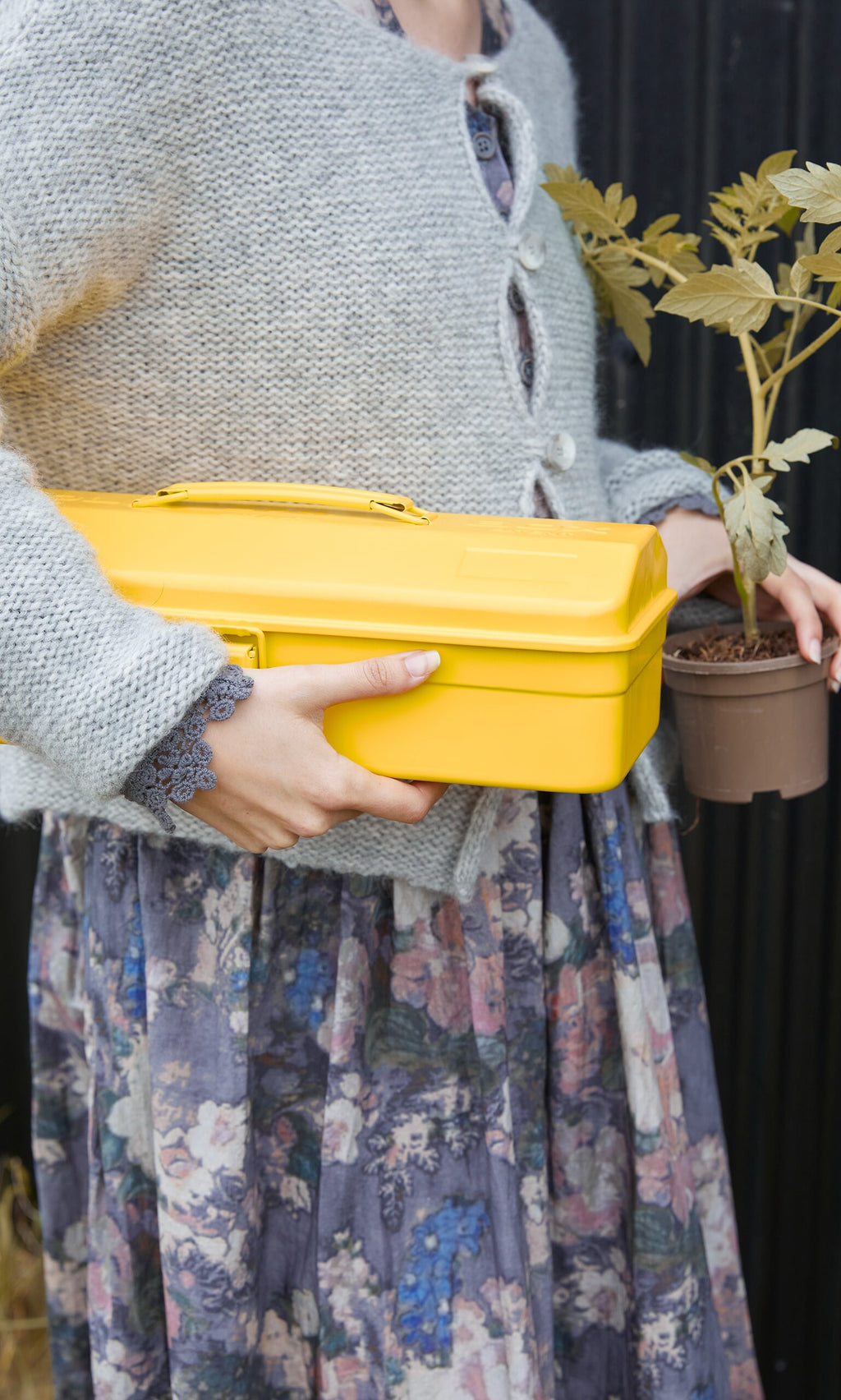 Plümo- Person in a grey cardigan and floral skirt holds a Yellow tool box, ideal for garden storage, plus a small potted plant.