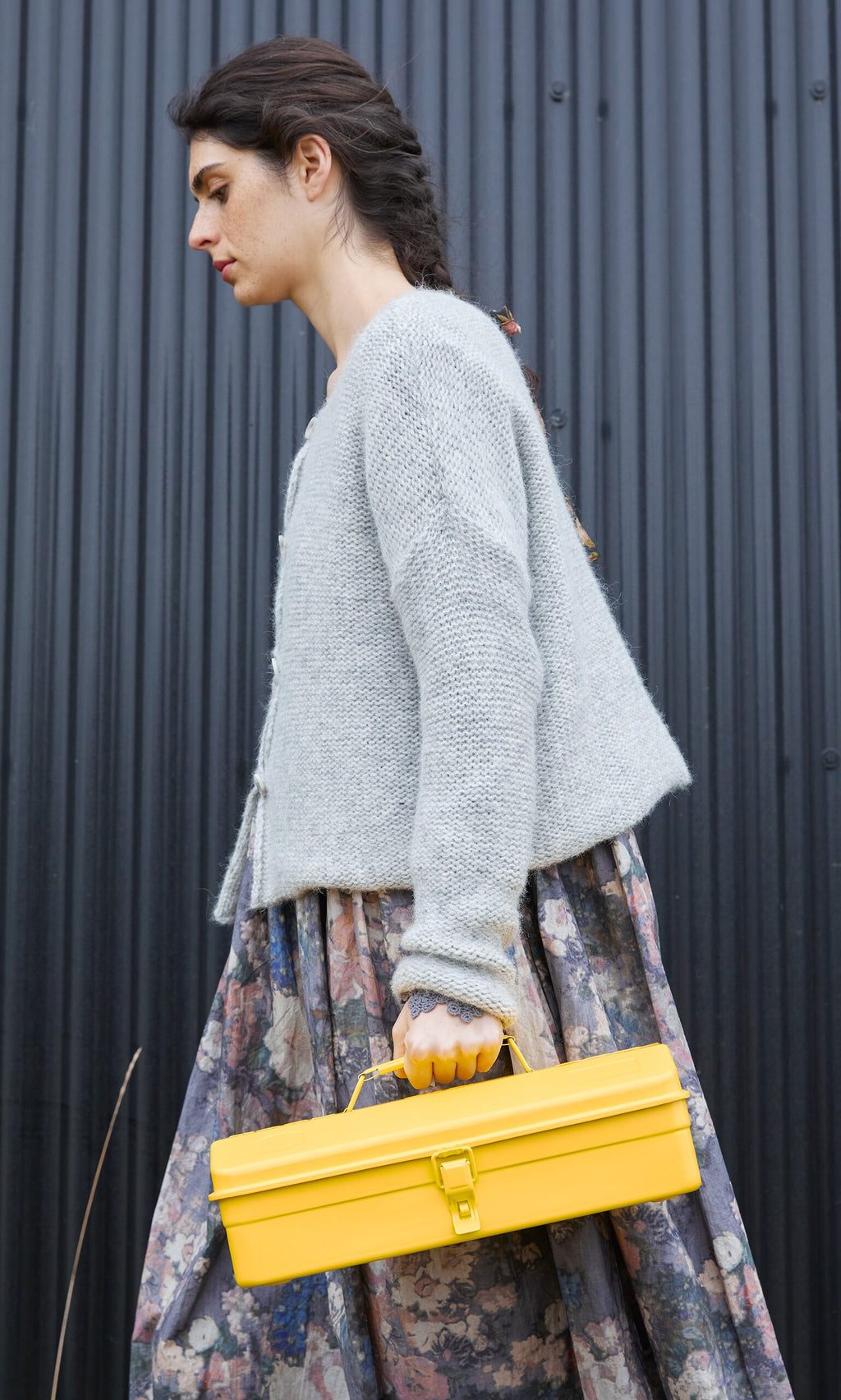 Plümo- Woman in a gray sweater carries a Yellow tool box for gardening, wearing a floral skirt by a black corrugated wall.