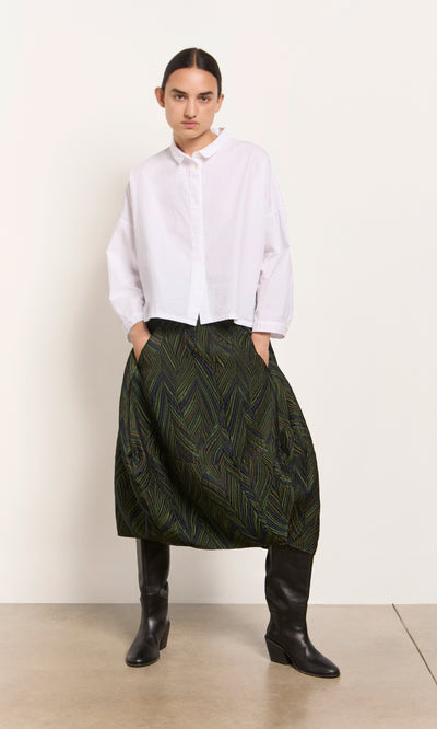 Plümo  Person in a white button-up shirt, Yona Skirt, and black boots stands against a plain off-white background.