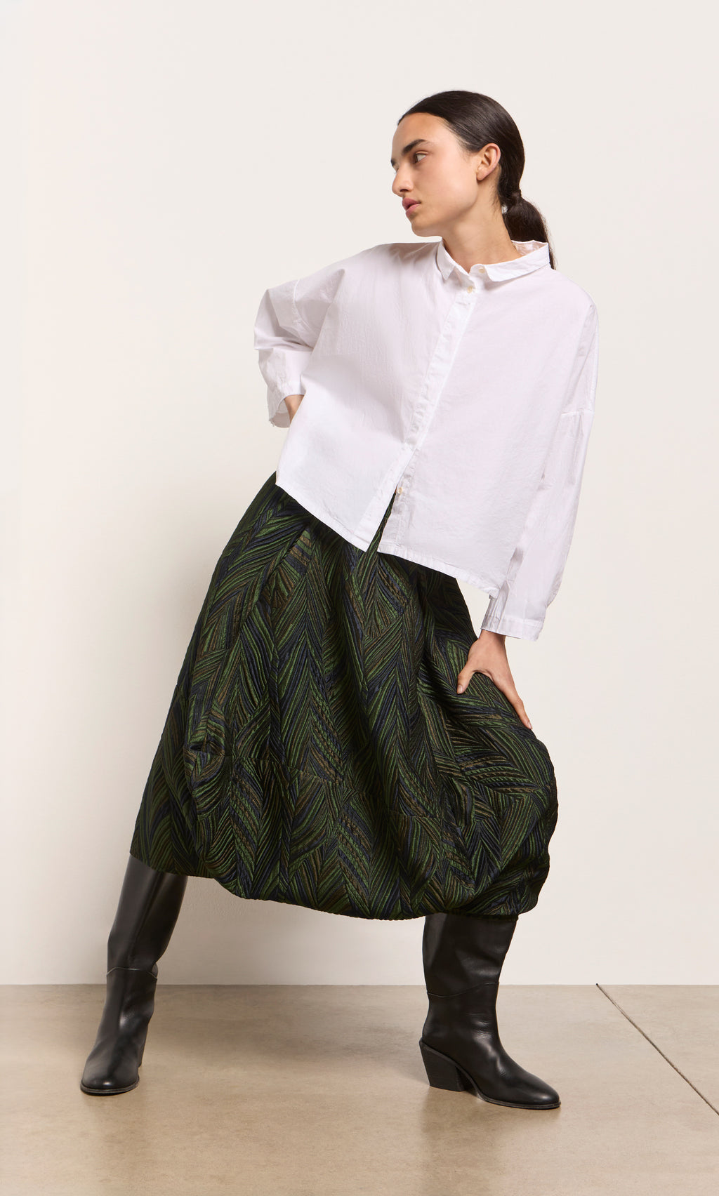 Plümo  Woman in a white shirt, Yona Skirt, and black boots poses against a plain light wall.