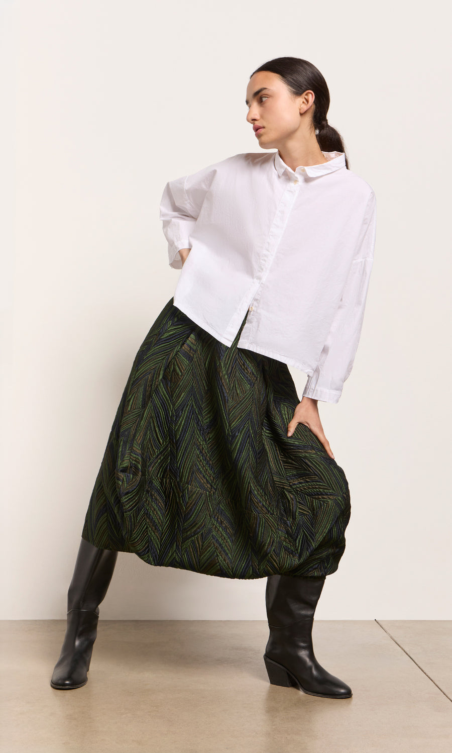 Plümo  Woman in a white shirt, Yona Skirt, and black boots poses against a plain light wall.