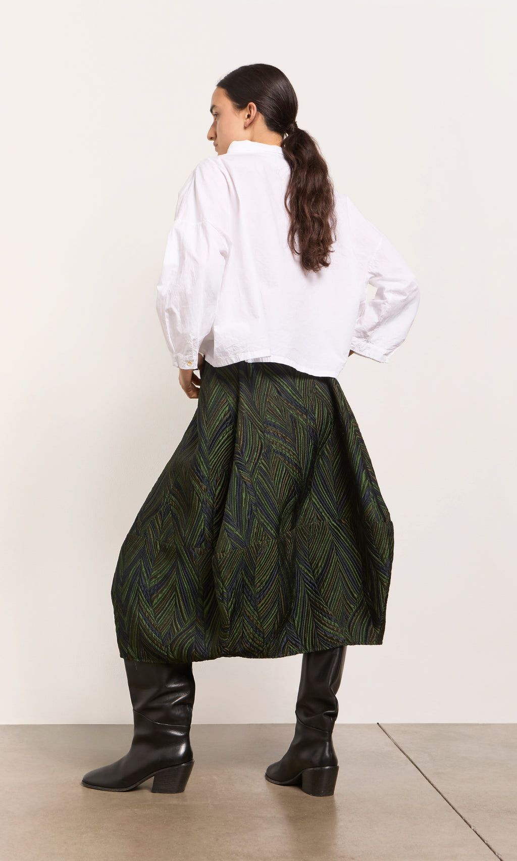 Plümo  Woman facing away in white blouse, Yona Skirt, and black knee-high boots on a plain background.