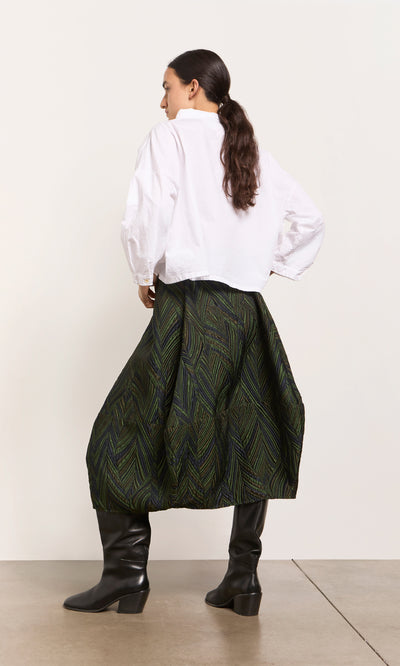 Plümo  Woman facing away in white blouse, Yona Skirt, and black knee-high boots on a plain background.