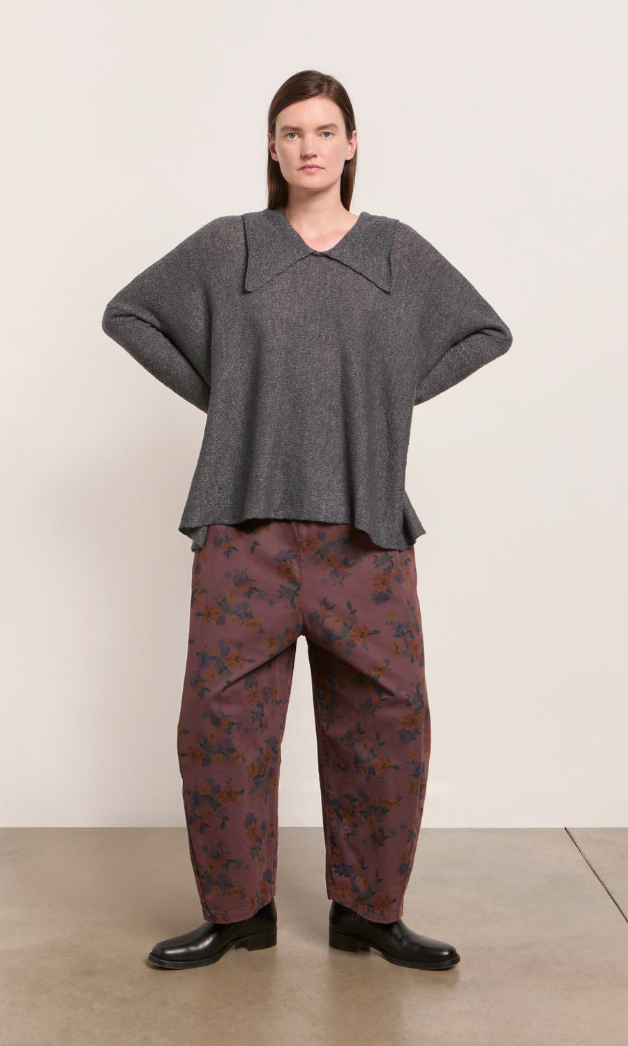 Plümo  Woman stands with hands on hips in Espel Pants, a gray sweater, and black shoes against a plain backdrop.