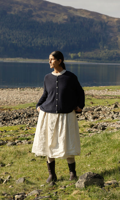 Plümo  A woman in the Alma Cardigan - Dark Navy stands on rocky grass by a lake, with hills and trees in the background.