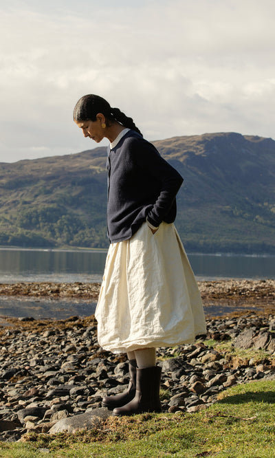 Plümo  Woman in the Alma Cardigan - Dark Navy and a cream skirt stands by a rocky lake shore with hills behind her.