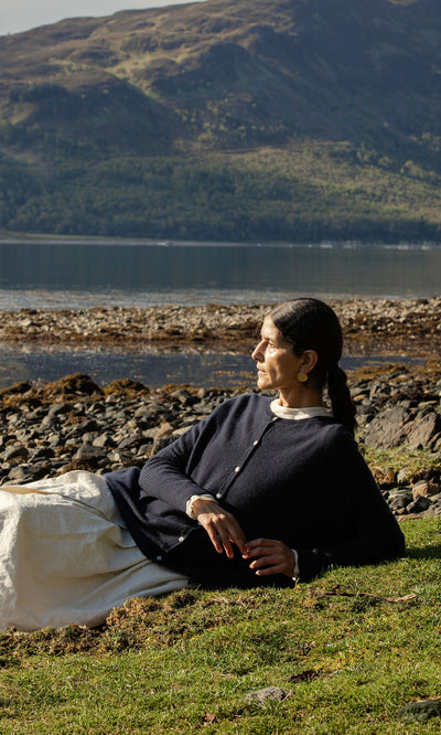 Plümo  Woman in Alma Cardigan - Dark Navy and white skirt lounges by a rocky lakeshore with mountains behind her.