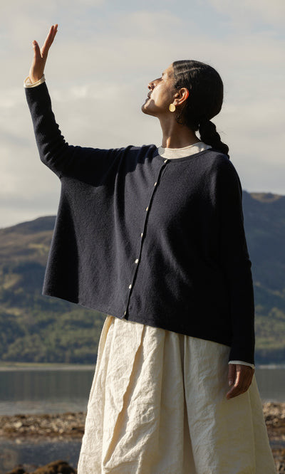 Plümo  Person in Alma Cardigan - Dark Navy raises hand by a lake, mountains forming a stunning backdrop.