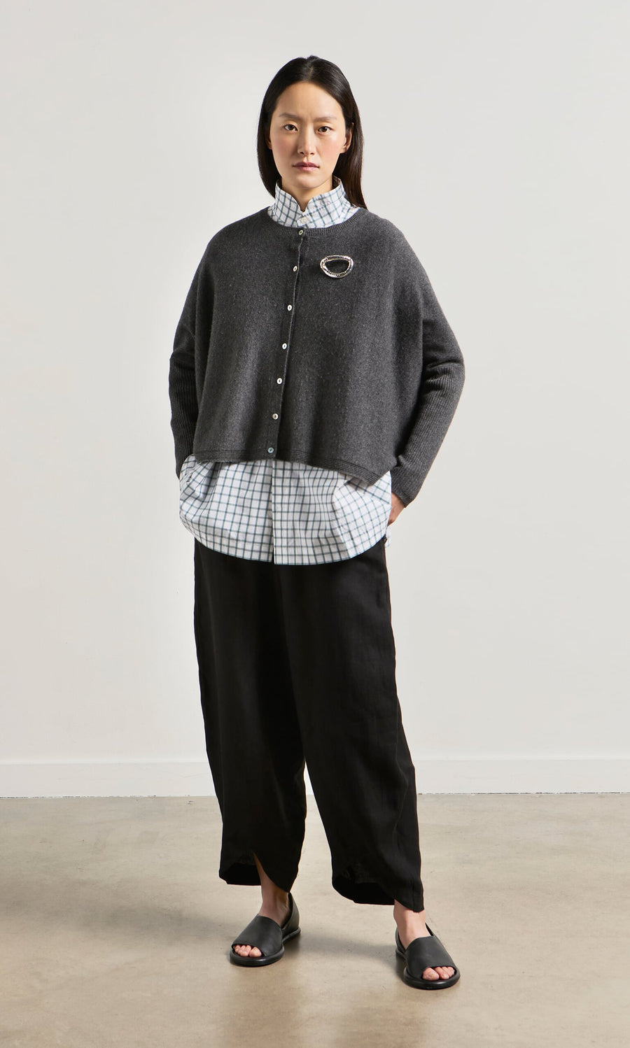Plümo- Woman standing against a plain wall, wearing the Alma Cardigan - Grey, a checkered shirt, black pants, and sandals.