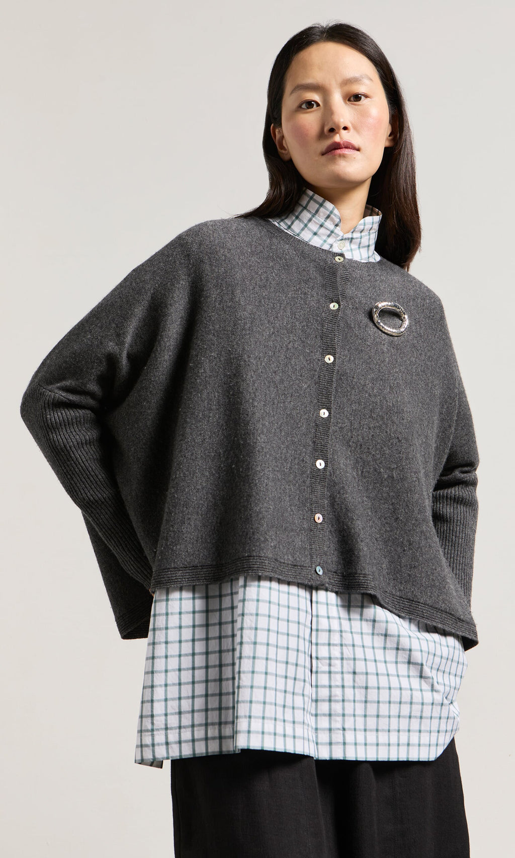Plümo- Woman wears the Alma Cardigan - Grey over a checkered shirt and black pants, standing against a plain background.