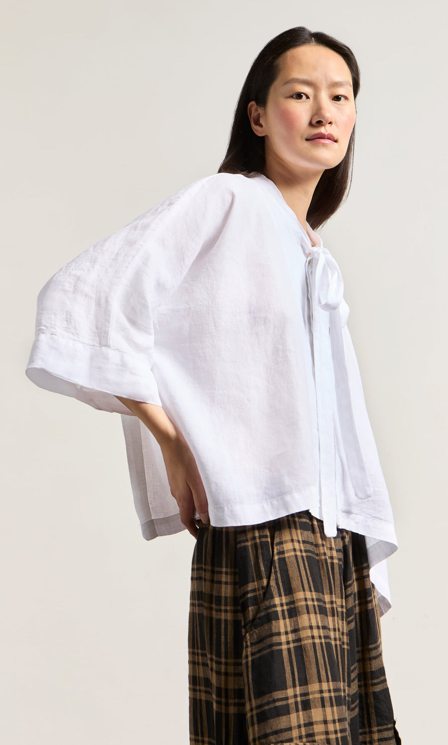 Plümo- Woman wears the Antoine Blouse - White with wide sleeves and ribbon tie, paired with a brown plaid skirt.