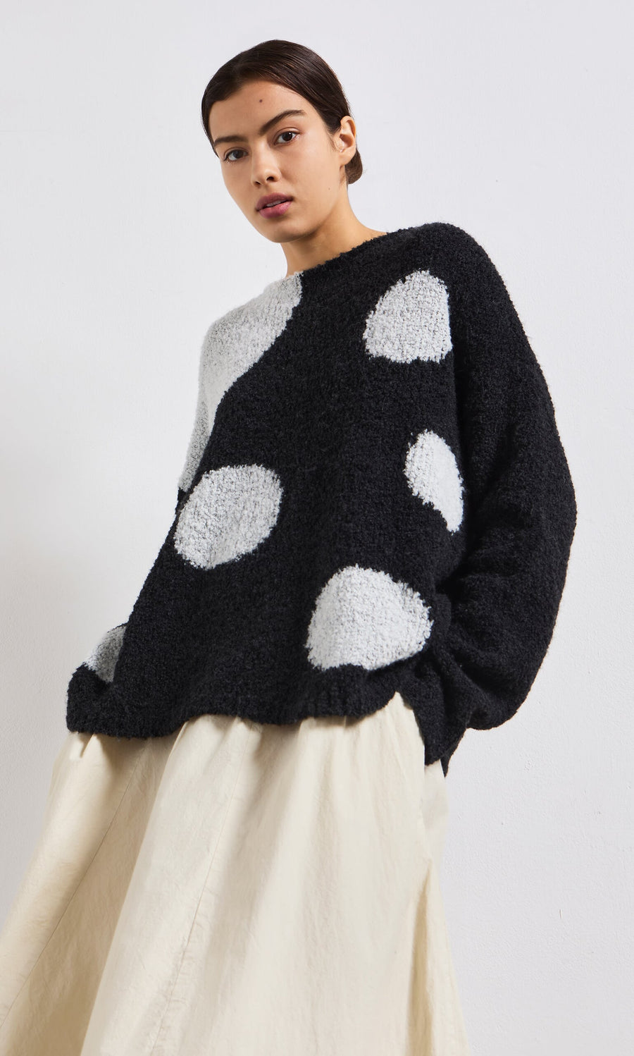 Plümo- Model wears the Antra Spot Jumper, a lightweight black and white polka dot knit, with a cream skirt against a white background.