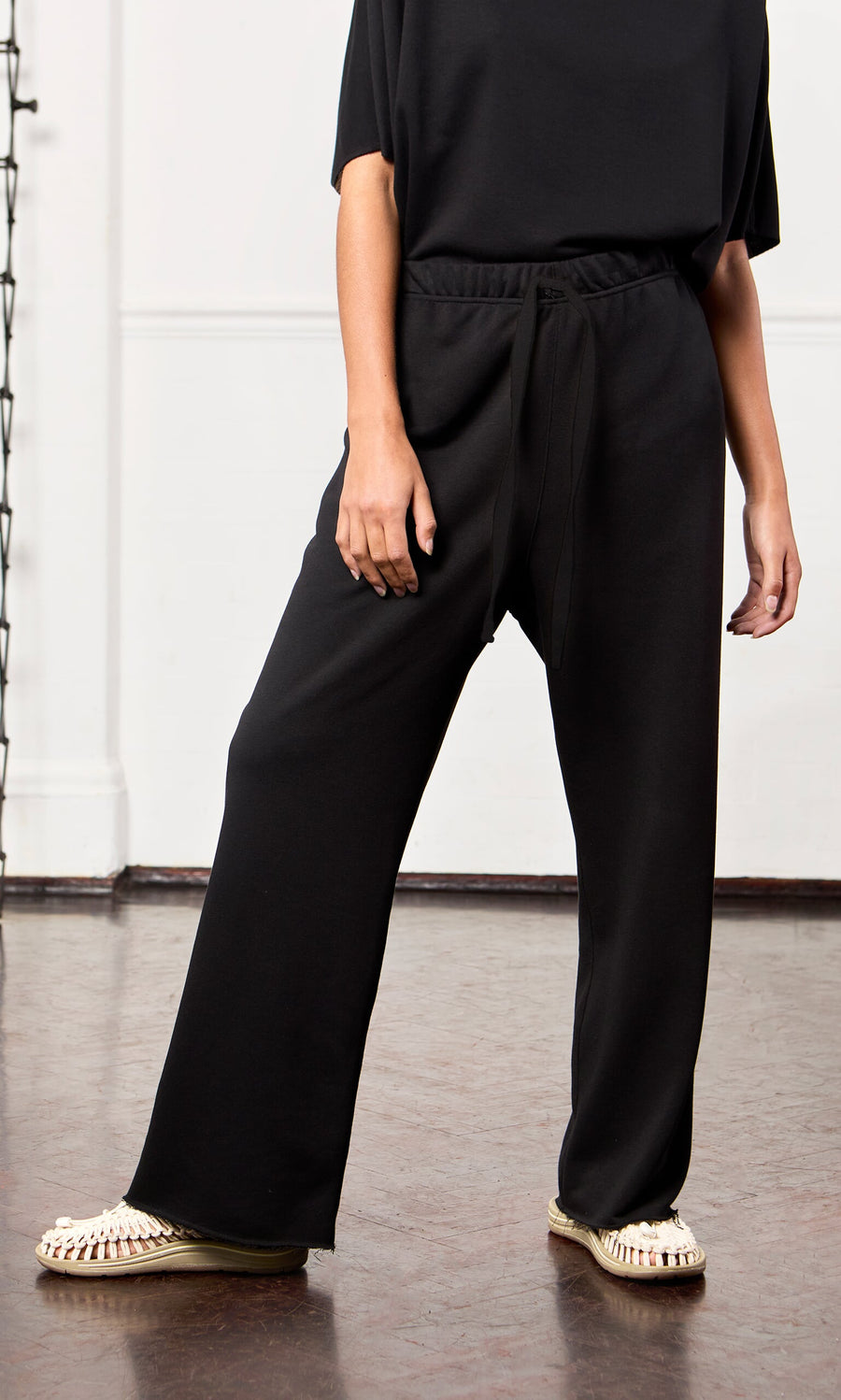 Plümo Person wearing Gabriella Pants—black cotton jersey lounge pants with elastic waist—plus a black top and white sandals indoors.