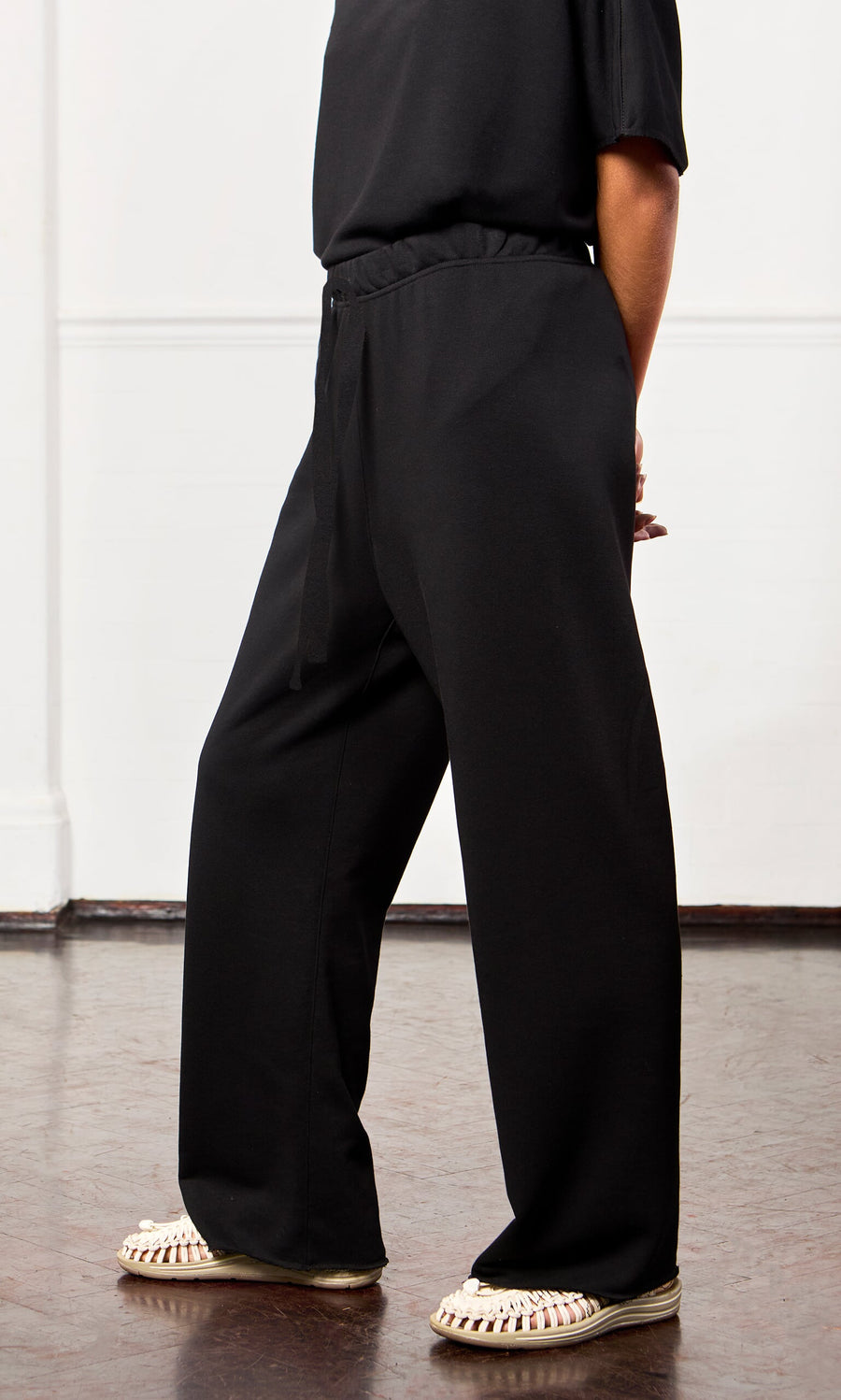 Plümo - Person indoors wearing Gabriella Pants with an elastic drawstring waist, black top, and white woven sandals.