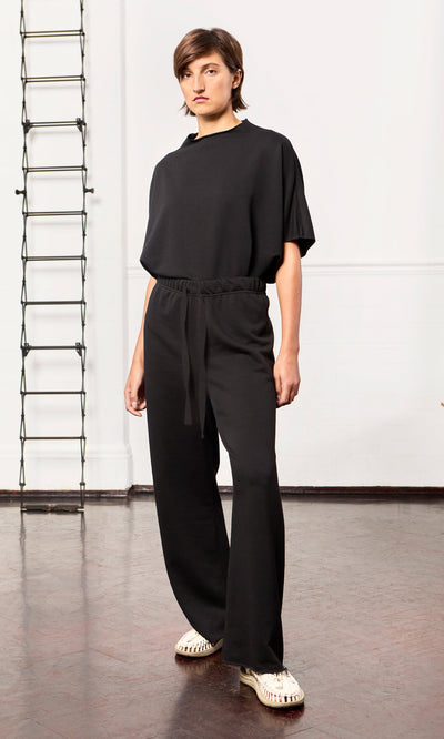 Plümo - Person in loose black Gabriella Pants and light shoes stands indoors on a brown floor with a ladder in the background.