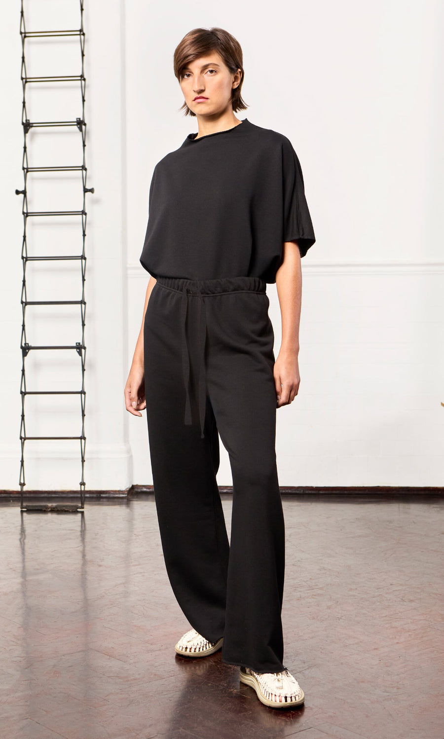 Plümo - Person in loose black Gabriella Pants and light shoes stands indoors on a brown floor with a ladder in the background.